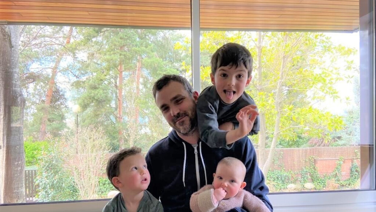 Iñaki Urgarte con sus tres  hijos: dos niñas y una niña. Están apoyados delante de un ventada que da a un jardín muy verde. Iñaki va con unos jeans y una chaqueta con cremallera de color azul. Unos de los hijos está apoyado en su espalda, mira a cámara sacando la lengua. Otro hijos está al lado izquierdo y se mira a su hermano. La niña, la más pequeña, va con un pijama body de color rosa y tiene una mano en la boca. 