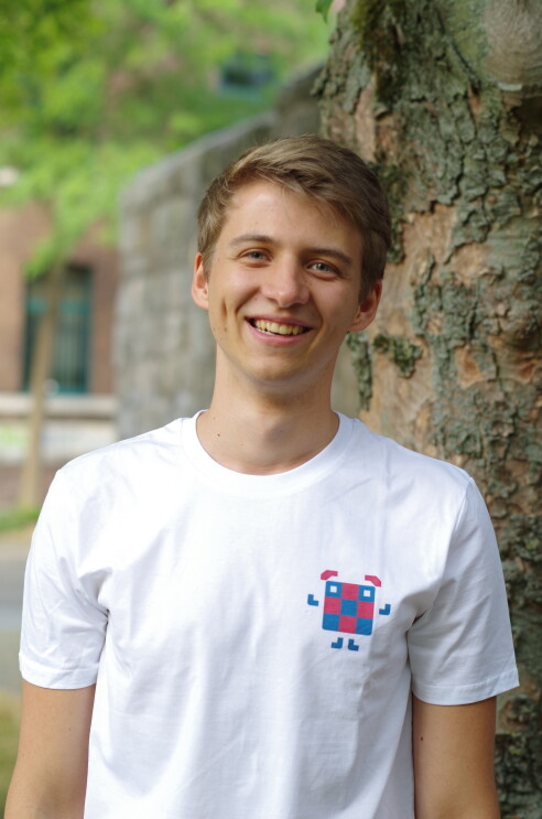 Eine junge mit weißem T-shirt und blonden Haaren steht vor einem Baum und lacht in die Kamera.