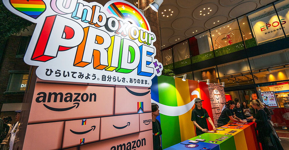 商業施設に設置されたイベントブース。「Unbox our PRIDE」と大きく書かれたボードの横に、レインボーカラーの衝立とカウンター。カウンターの内側に揃いのTシャツを着たスタッフが立っている。
