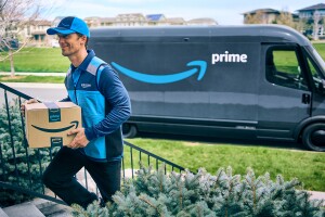 Amazon Prime delivery driver walks up the steps to deliver a package with his Prime delivery van in the background.