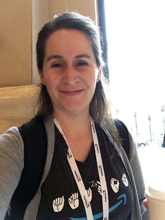 A woman stands outside, wearing a backpack, lanyard, and t-shirt with the Amazon logo in American Sign Language. 