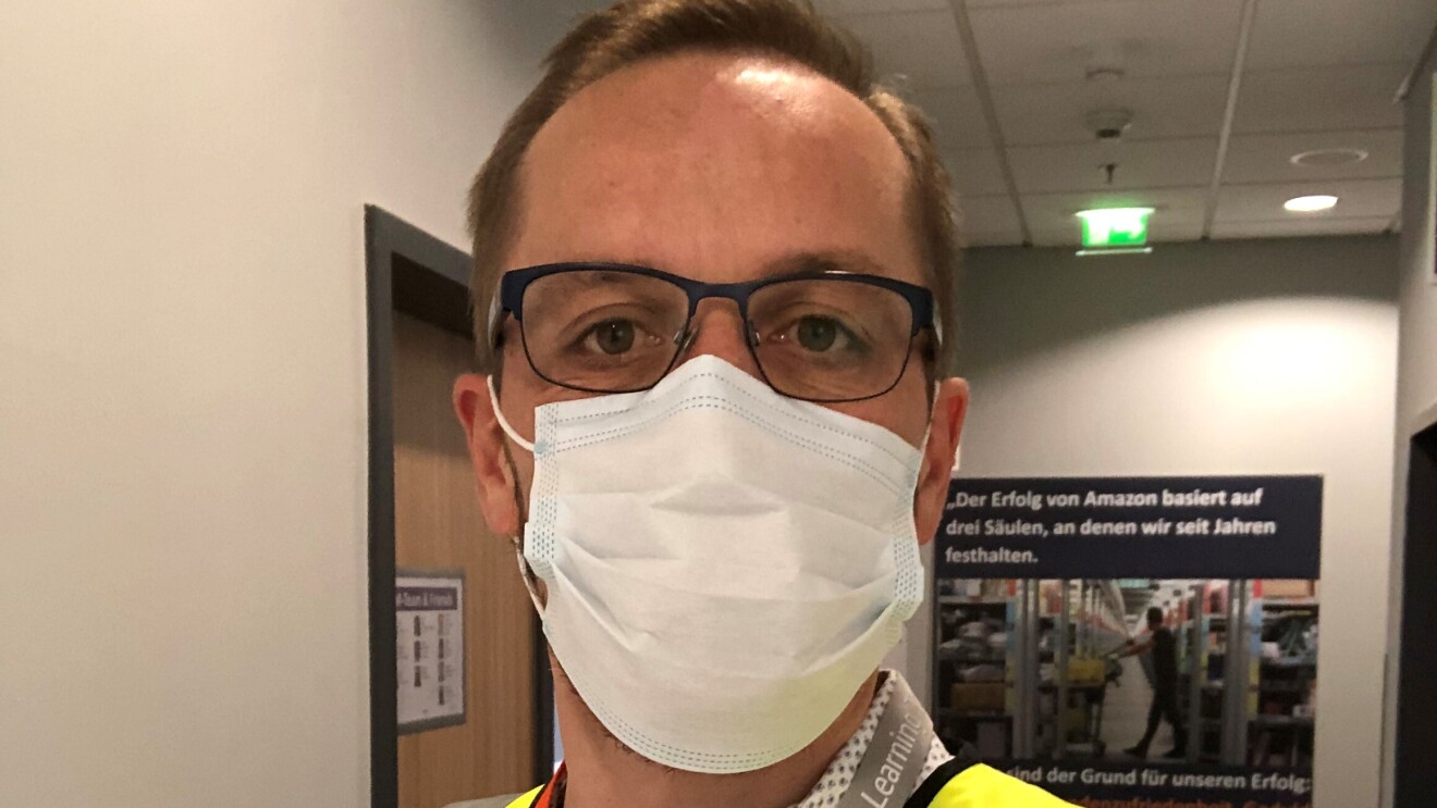 Ein Mann mit blonden Haaren, Brille und Sicherheitsweste vor einer Packstation im Logistikzentrum