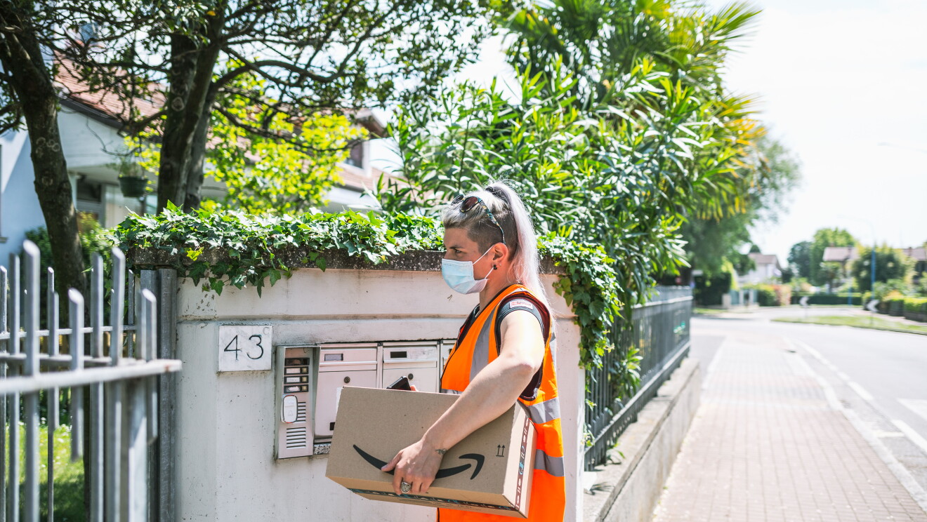 Elisabetta si trova di fronte al cancello di un'abitazione. Porta un pacco Amazon sotto il braccio sinistro, è girata di lato. Indossa un giubbino da lavoro arancione ed è ripresa a busto intero. Sullo sfondo si intravede un paese. 
