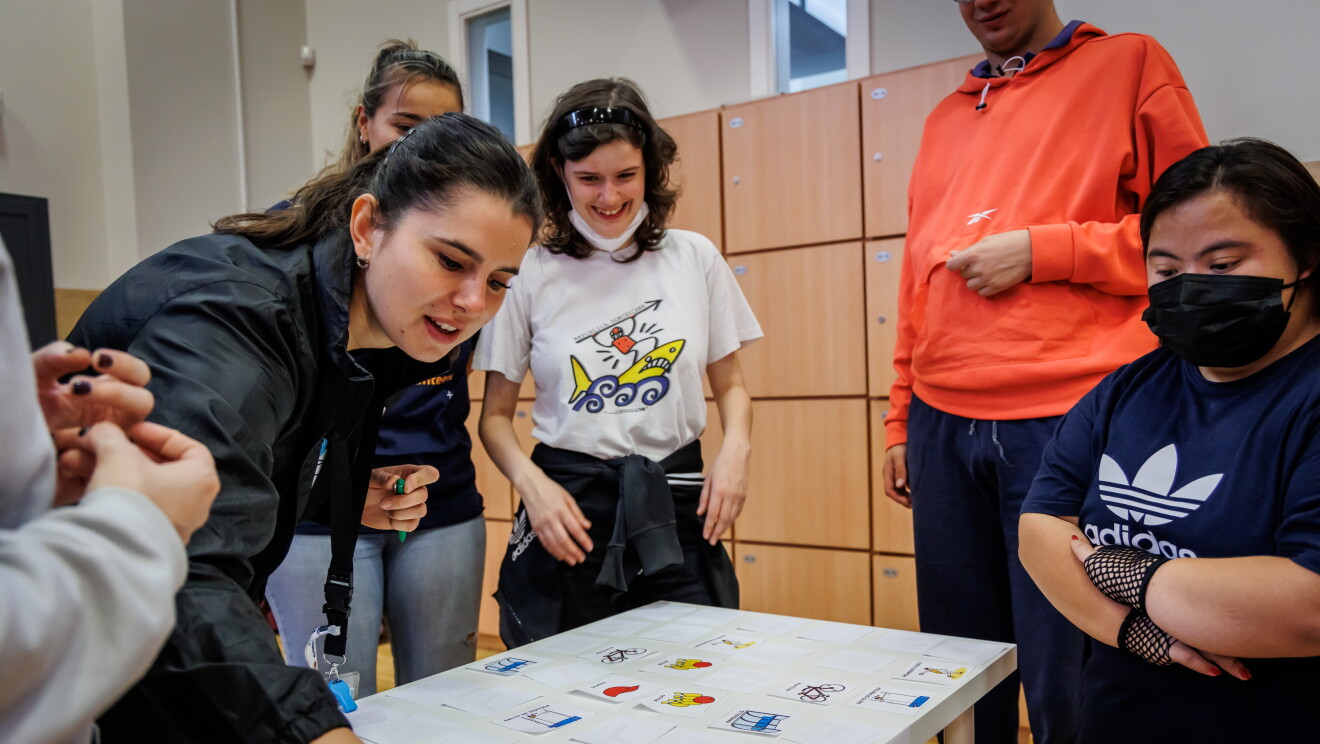 En las instalaciones de Down Madrid en Madrid aparecen 5 personas, 2 trabajadoras de Amazon y tres participantes de la fundación. Los trabajadores de Amazon van con la misma camiseta azul y el badge colgado y los de Down Madrid con deportivas y ropa cómoda. Una de las trabajoras de Amazon está participando en la gincana colocando un dibujo de un a pelota de tennis sobre una mesa. 