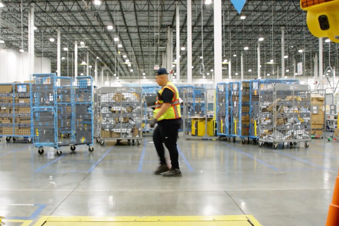 Charles wears a yellow safety vest and walks through an Amazon fulfillment center.