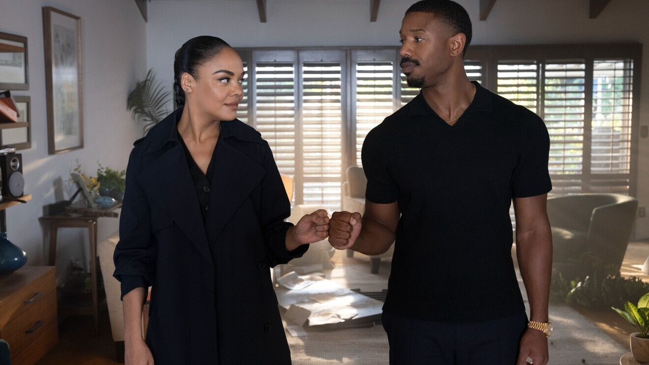 Michael B. Jordan and Tessa Thompson are both dressed in black and fist bump while standing next to one another in a home.