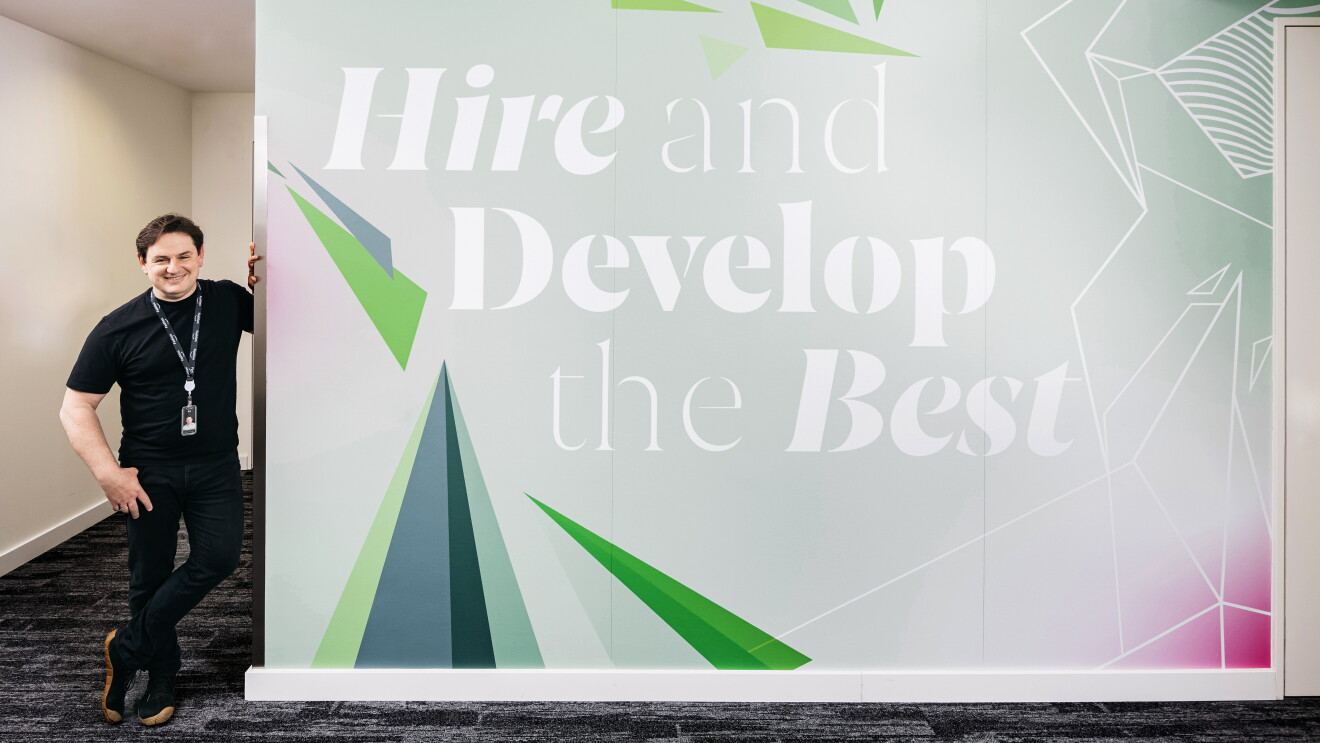 Kai Rühl (ein Mann in schwarzen Jeans, Sneakers und T-Shirt, mit braunen Haaren) lehnt an einer Wand in einem Amazon Bürogebäude auf der eines der Leadership Principles illustriert wurde: Hire and Develop the Best.