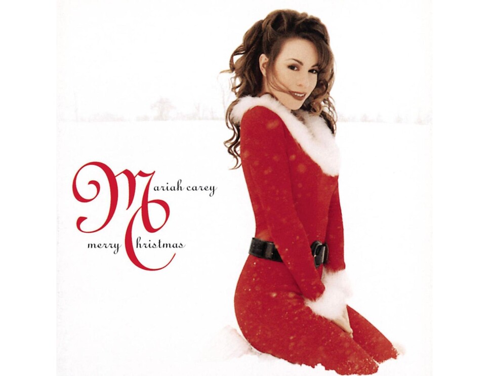 Mariah Carey wears a red dress with furry trim at the collar and cuff, and a thick, black belt. her outfit is reminscent of Santa's traditional Christmas look. She's kneeling on fresh snow, and looking to her left, at the camera. Behind her, in the distance are the hint of trees. 