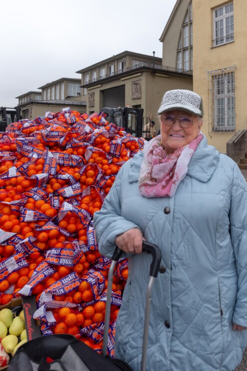 Eine ältere Dame mit Brille und Schildmütze im hellblauen Steppmantel steht vor einem großen Berg von Mandarinen und Orangen.