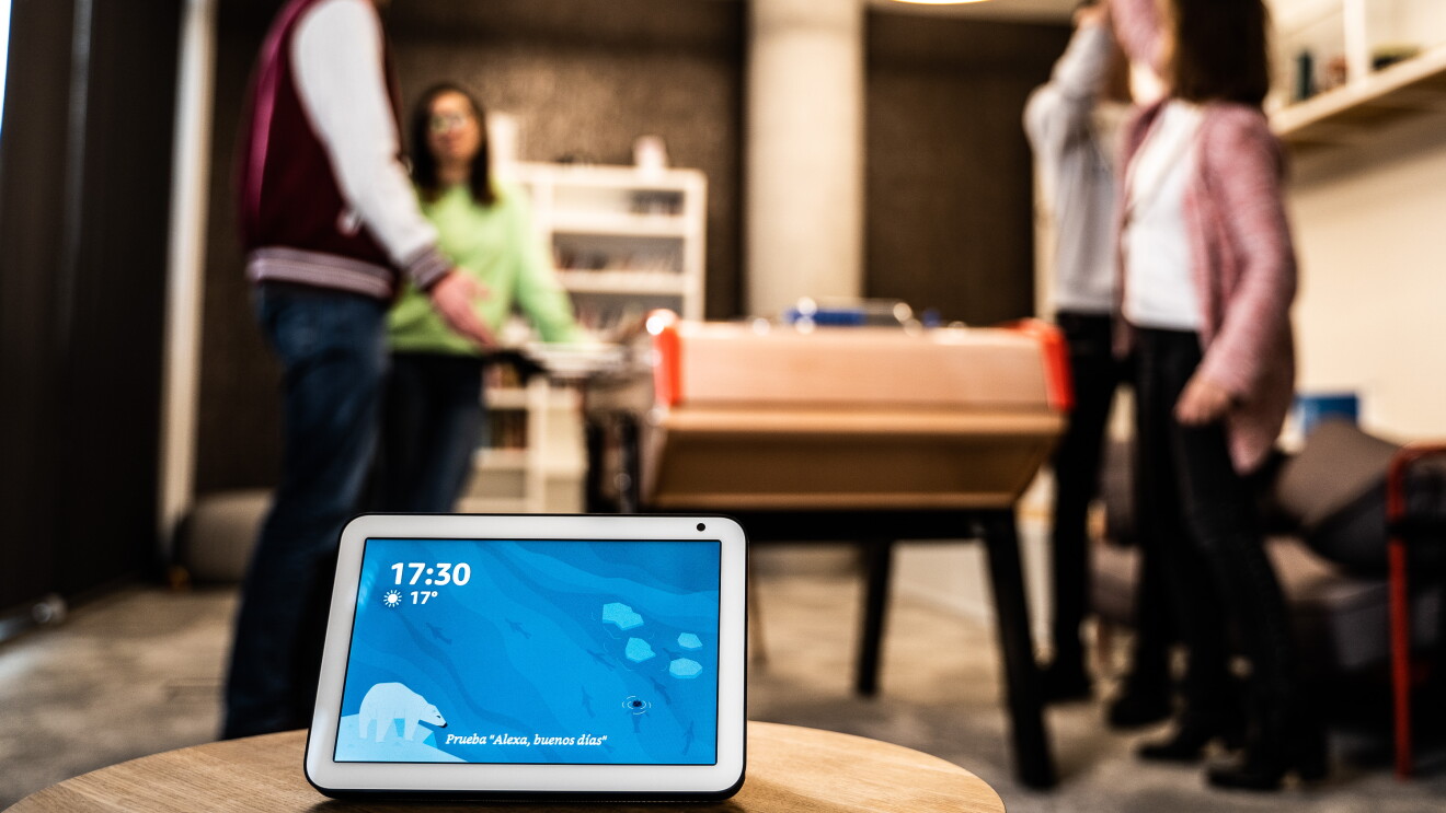 El dispositivo Echo Show en primer plano con la pantalla de color azul la hora y la temperatura. EL dispositivo se encuentra encima de una mesa. De fondo se ven cuatro personas jugando al futbolín. Dos chicos y dos chicas. Las parejas son mixtas y una de ellas se está celebrando que ha marcado un gol. 