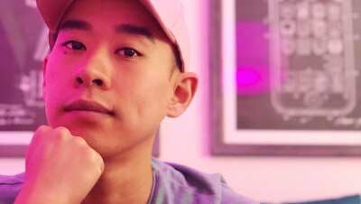 一名男子在粉色灯光下微笑拍照的大头照。他戴着一顶白色的棒球帽,下巴托在拳头上。