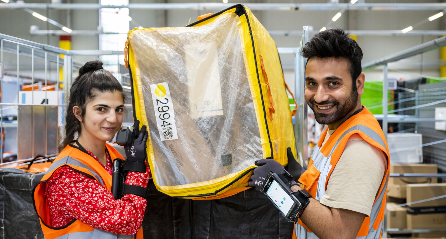 Zwei Amazon Mitarbeiter:innen (eine weibliche Mitarbeiter und eine männliche) halten einen gelben Transportbehälter in den Händen.