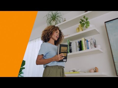 An image of a woman holding a book while talking to Alexa. There is an orange stripe on the left side.