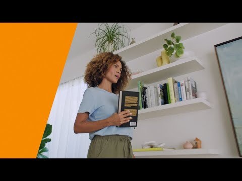 An image of a woman holding a book while talking to Alexa. There is an orange stripe on the left side. 