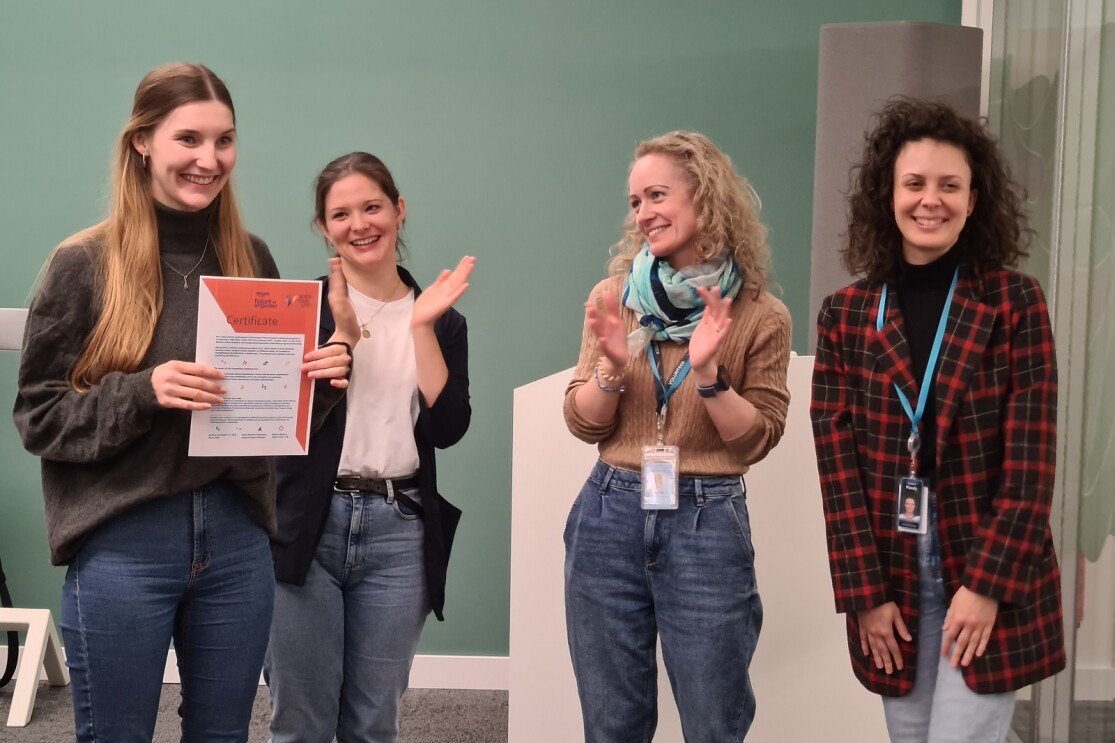 Teilnehmer:in empfängt ein Zertifikat bei der Abschlussfeier der ersten Runde des Mentoring-Programms. Dieses wird durch vier Personen übergeben. Alle angezeigten Personen lächeln und klatschen.