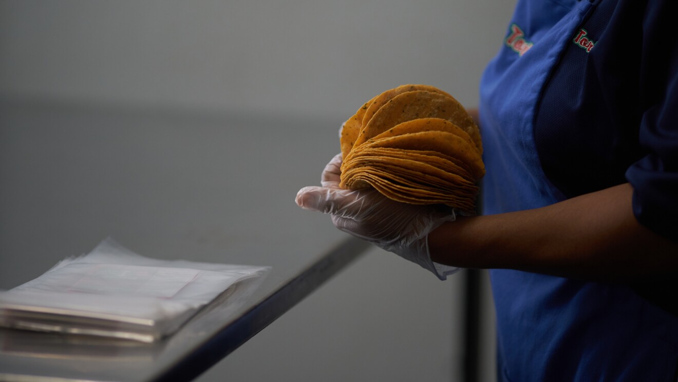 Primer plano de las manos de una mujer que va con guantes transparentes y agarra tortillas. Va vestida con un delantal azul y una camisa azul oscuro. Está de pie delante una mesa metálica y unas bolsas de plástico transparente.