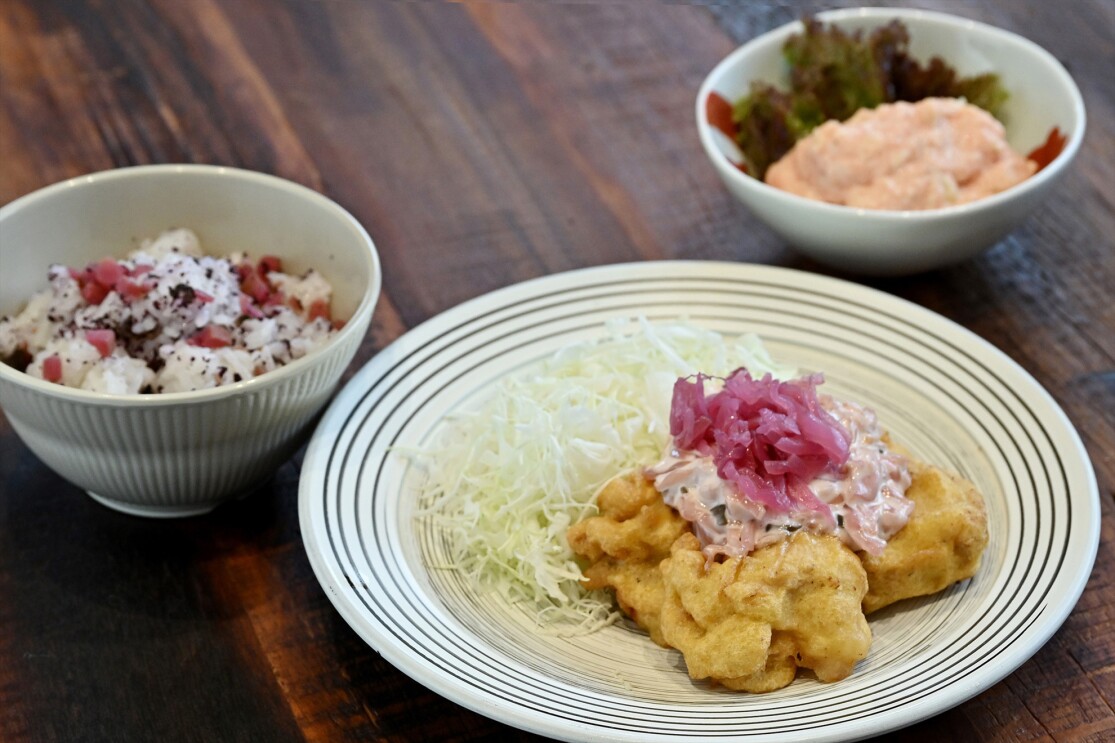 チキン南蛮に赤いシソ漬けが載った皿とお茶碗に入ったシソのふりかけがかけられた白いご飯、ピンク色のポテトサラダが入った小鉢