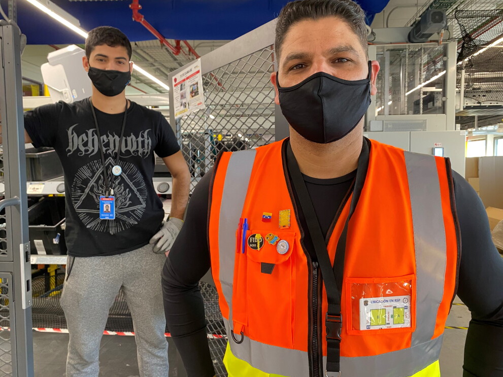 Daniel Fonseca y Diego Fonseca en el centro logístico de Amazon en El Prat de Llobregat (Barcelona).  De fondo unas cajas de color negro y el techo con conductos de color azul. En primer plano Daniel con una camiseta térmica estrecha negra de manga larga y un chaleco naranja. Mascarilla de color negro, pelo negro corto y mirando a cámara. En segundo plano está Diego, con un pantalón de deporte una camiseta negra de manga corta. Lleva guantes y la mano izauierda la apoya en la cintura y la derecha en una puerta metálica. Mascarilla negra.   