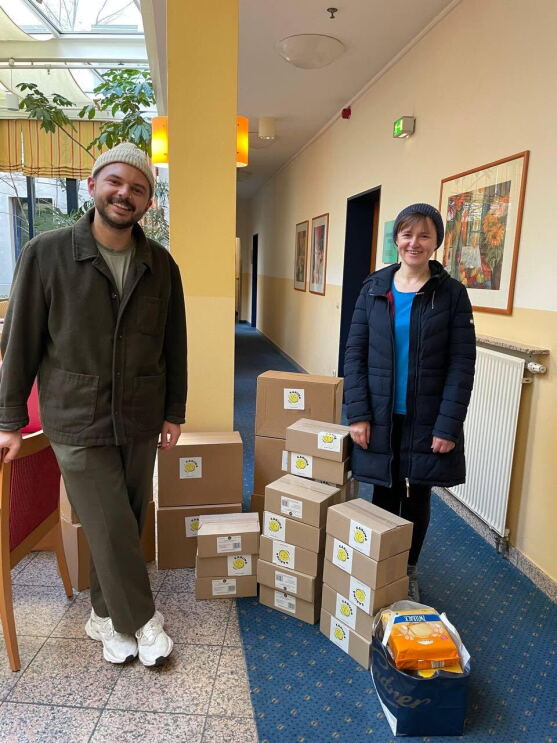 Zwei freiwillige Helfer von Amazon stehen neben diversen Kisten mit Sachspenden wie Powerbanks, Lebensmitteln und Babyartikeln für die gemeinnützige Organisation KARUNA eG.