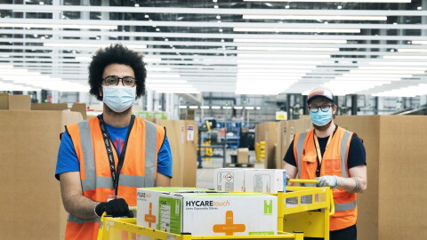 Amazon employees in a fulfilment centre packing medical supplies ready for the government into boxes. They are wearing hi-vis jackets and face masks. 