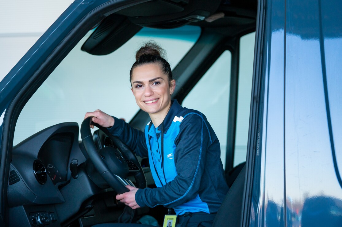 Eine junge Frau in blauer  Amazon Lieferuniform sitzt am Steuer eines Lieferwagens, sie blickt lächelnd in die Kamera.