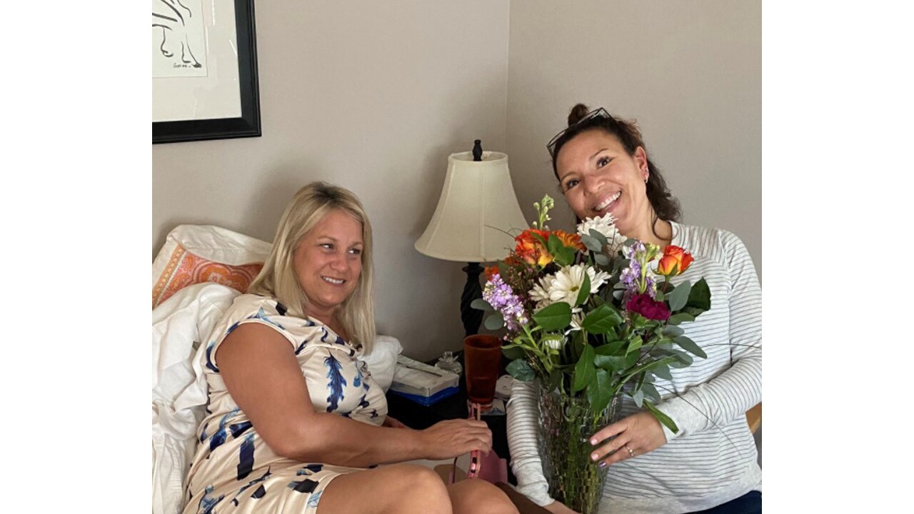 An image of two women smiling for a photo while sitting in a living room. One woman is holding flowers. 