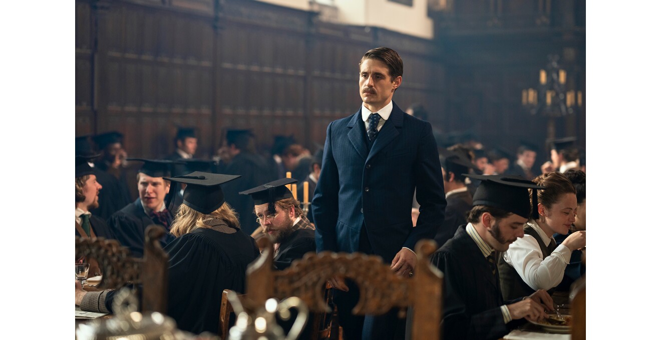 四角い学帽と黒いガウンを着用した古風な学生たちが大勢テーブルについて食事をしている食堂。口髭を生やしたスーツの男性がひとりだけ立っている。