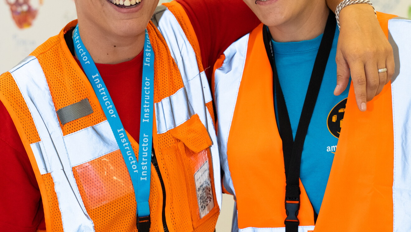 María García y Susana Torres fueron dos de las afortunadas que disfrutaron del desfile del Día del orgullo LGTBI desde la carroza de glamazon. la dos visten con los chalecos naranjas de Amazon y miran a cámara sonriendo. Tienen el pelo corto y pendientes. María abraza a su pareja del cuello. 