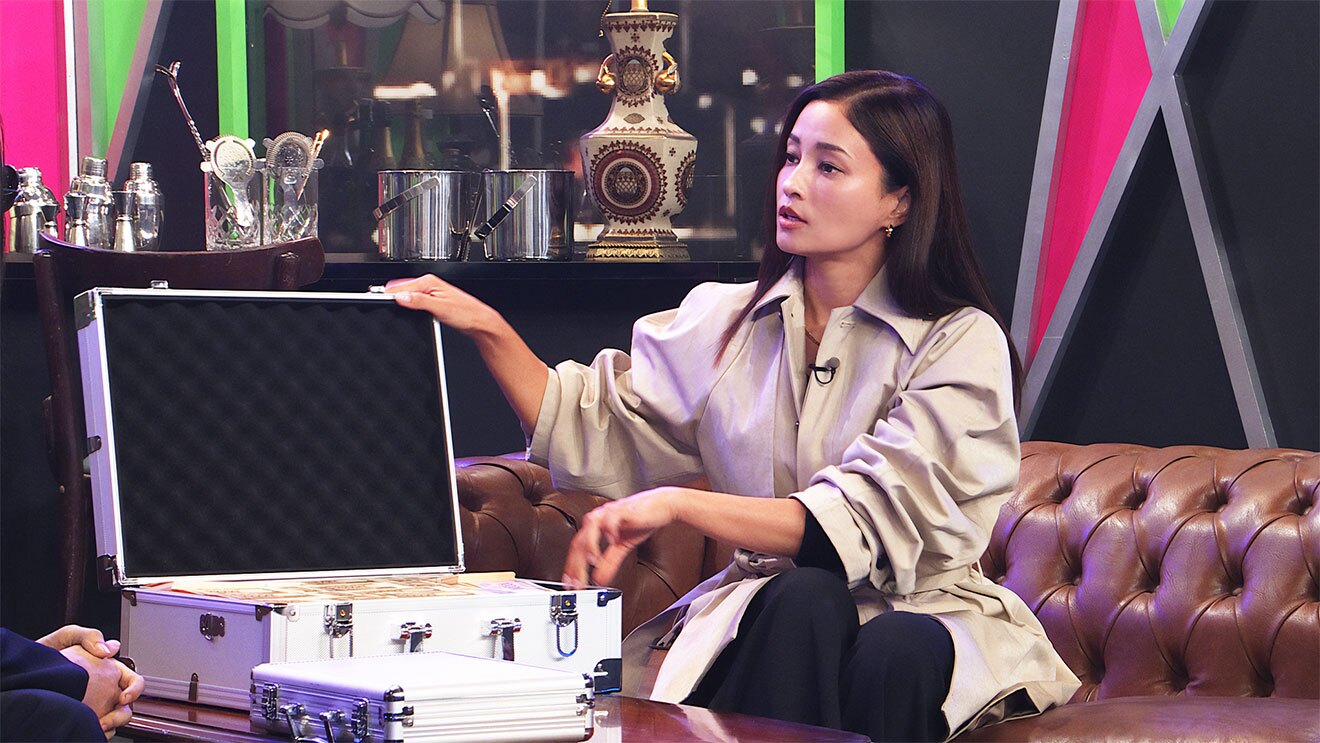 黒い壁の一部が黄緑やピンクに塗られたセットを背景に、茶色のソファに座る女性。ベージュ色のシャツに黒いパンツで、テーブルにのせたアタッシュケースを開いて蓋に右手をかけている。