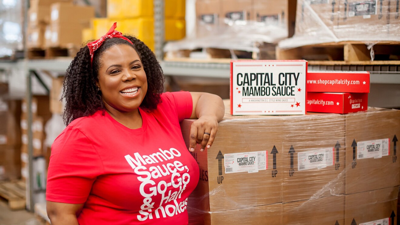 一名女子穿着写着“Capitol City Mumbo Sauce”的衬衫微笑着拍照。在一个满是箱子的仓库里,她靠在一箱箱子上。