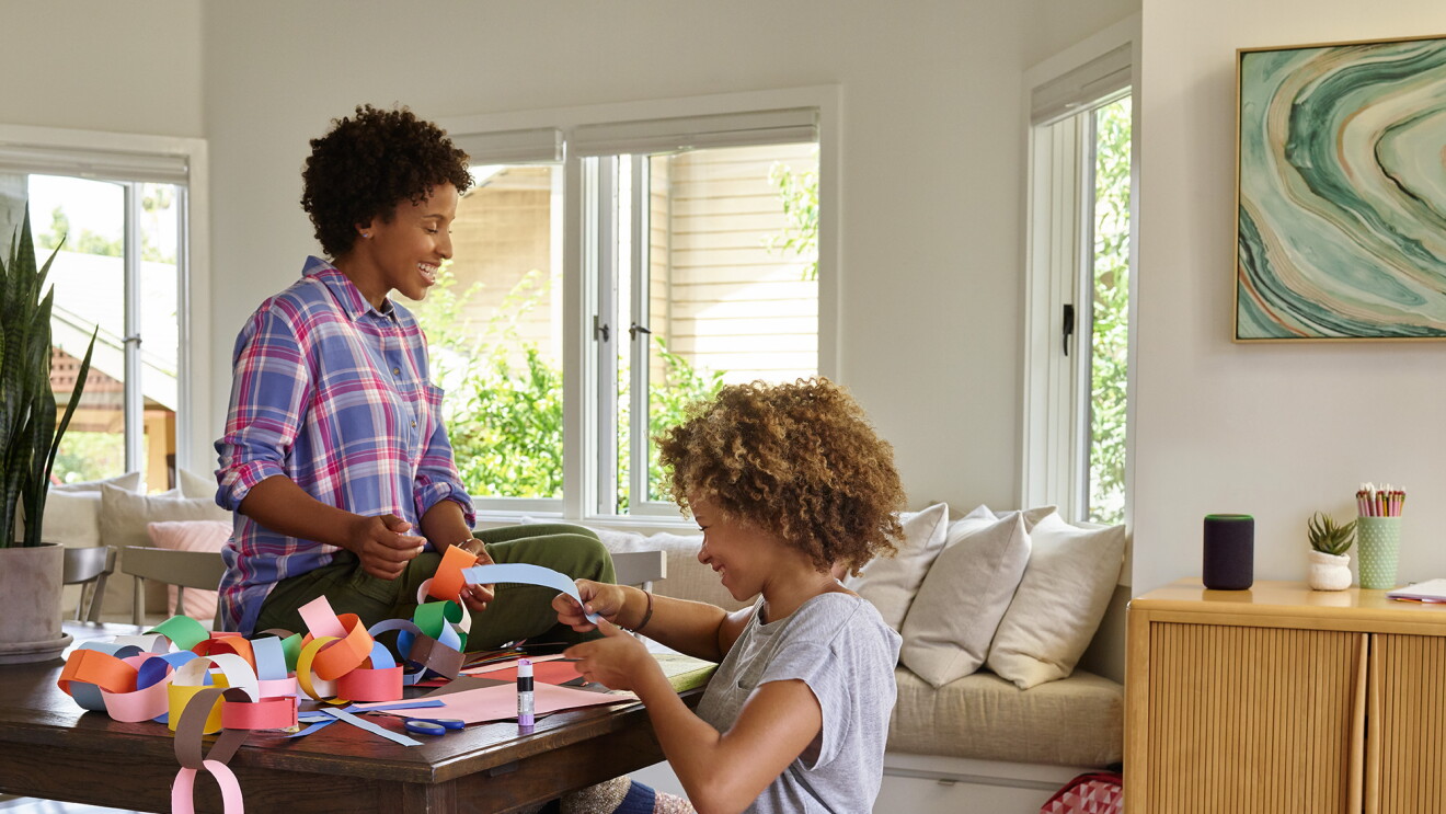 A mother sits on a dining room table as she and her daughter laugh, while making a paper chain. Behind them, Amazon Echo is on a console table. 