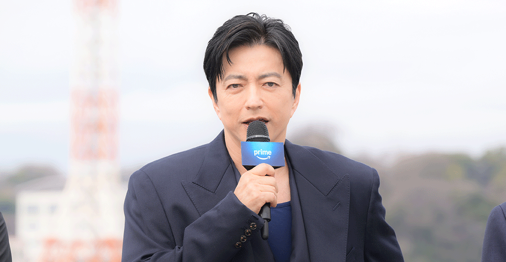 日中の屋外で、紺色のジャケットを着てマイクを持った男性。口にマイクを当て話している。背景に白と赤の電波塔のようなものが見える。