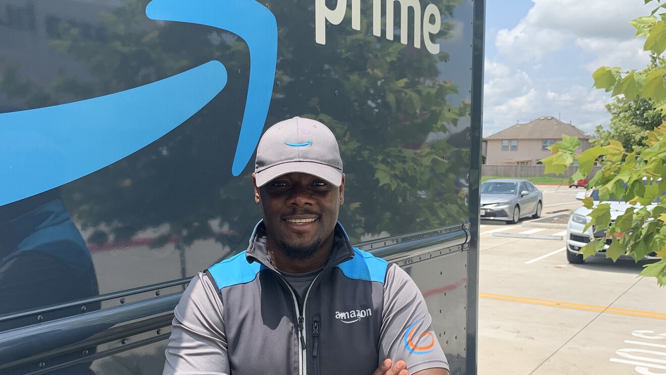 一名男子站在一辆亚马逊送货卡车前,卡车侧面写着亚马逊的标志和“Prime”字样。他戴着亚马逊品牌的帽子和背心,双手交叉在胸前微笑着。