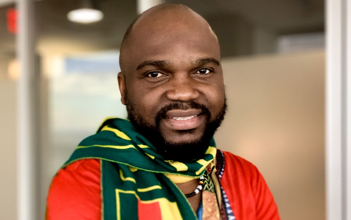 An image of a man smiling for a headshot photo. He is wearing a bright red shirt and a green and yellow scarf.