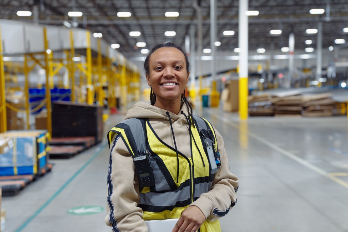 Image of employee Jessica standing in a fulfillment center holding her laptop and smiling.