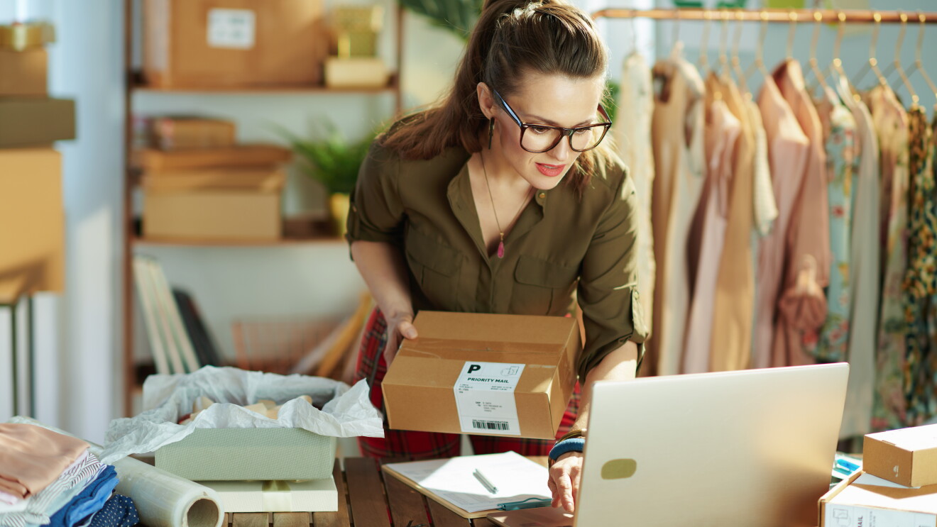 Eine Frau mit Brille steht vor einem Laptop und hat ein Paket in der Hand.