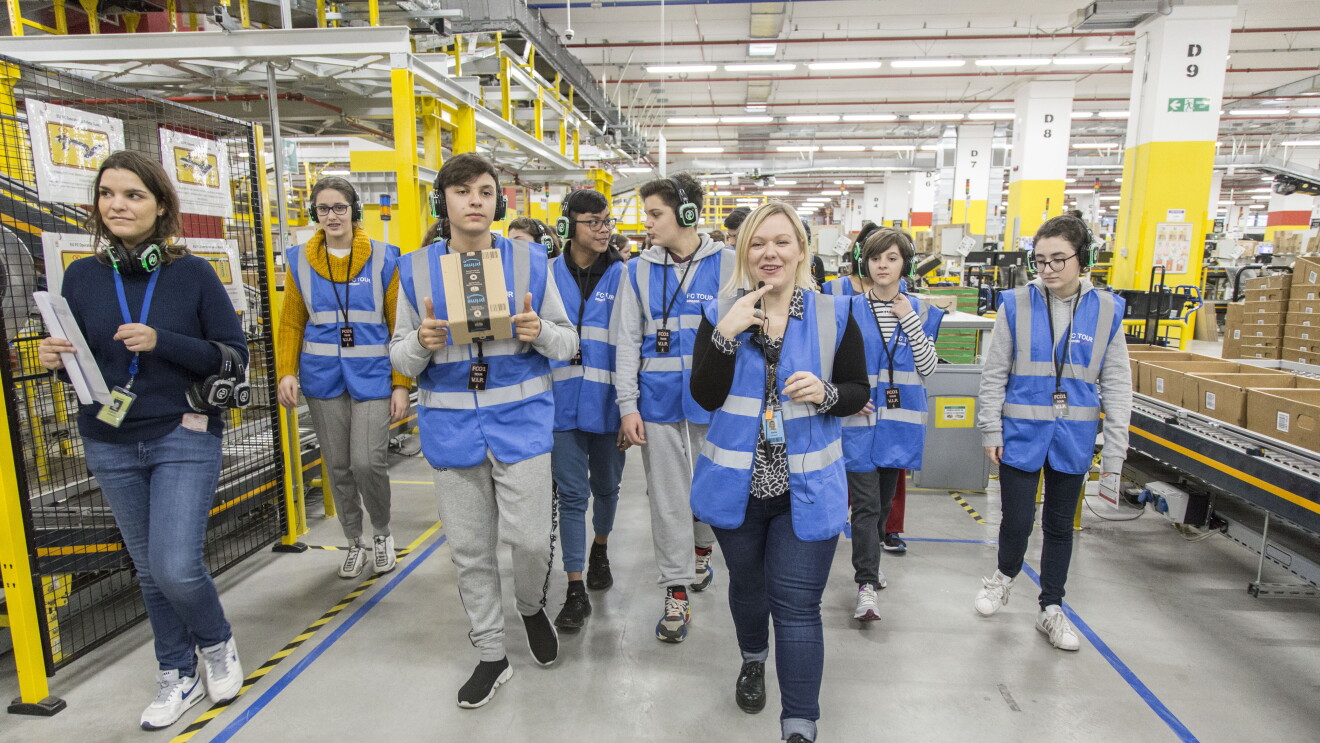 Una foto di Daria Banach, Tour Lead nel centro di distribuzione Amazon di Passo Corese, mentre accompagna i ragazzi di una scolaresca in un tour del magazzino. Daria parla al microfono e i ragazzi sono muniti di cuffie.
