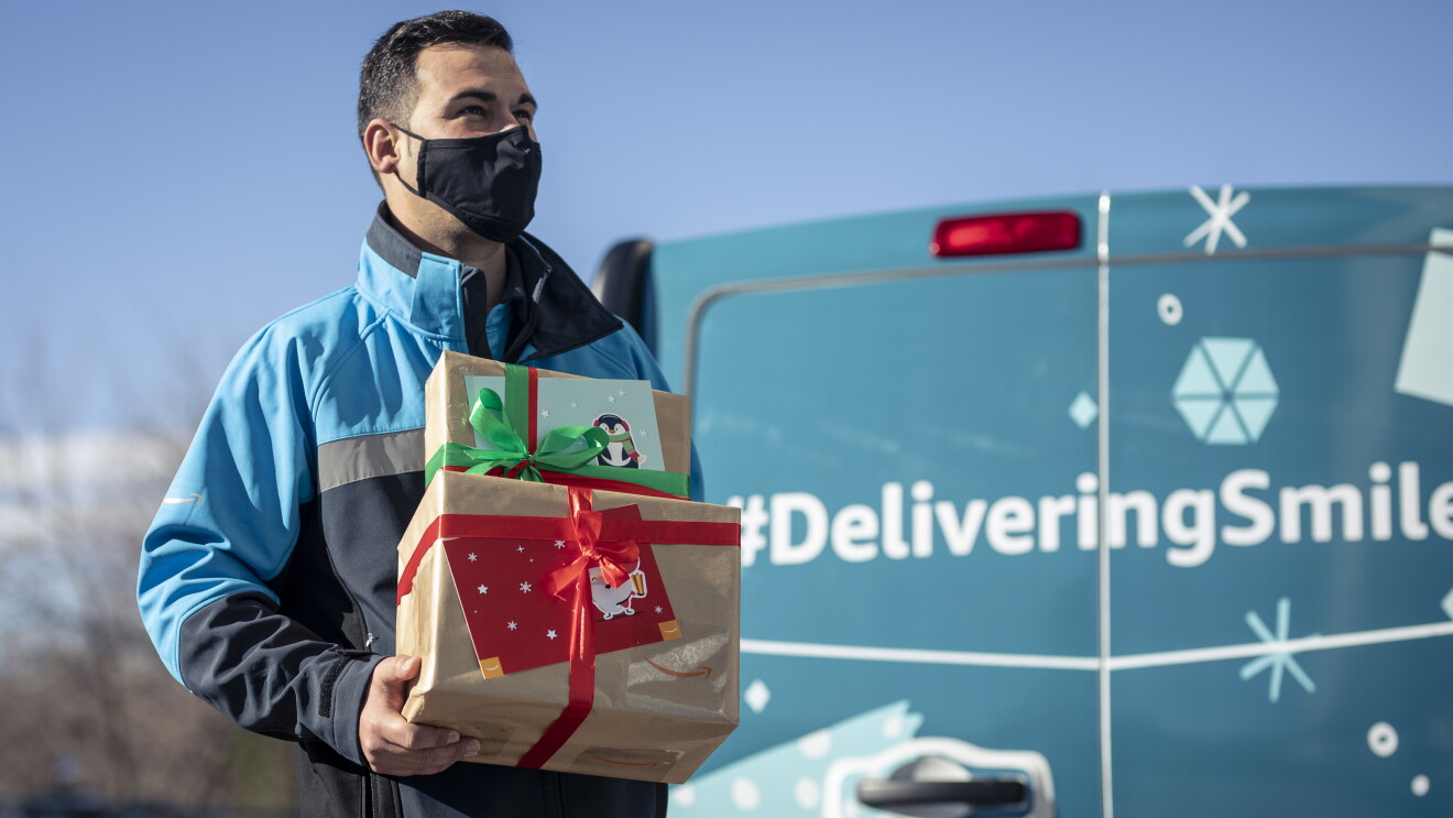 Donación al Centro de día de personas sin hogar Casablanca de Alcobendas.  A la parte derecha de la foto y en segunda plano, una furgoneta de Amazon de color azul-verde. Se ve solo la parte trasera y tiene la etiqueta impresa de DeliveringSmiles. En primer plano un repartidor de Amazon con pelo negro corto, mascarilla negra y un anorac de color azul claro, azul marino y gris. Carga dos cajas de Amazon envueltas en papel marrón con un lazo una rojo y la otra verde y dos tarjetas regalo. Mira al horizonte y se le imagina una sonrisa en el rostro. 