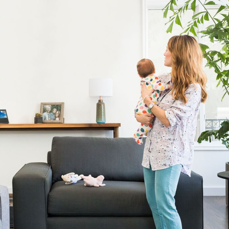 A mom holds her baby as she looks across the living room at a Echo Show device. 