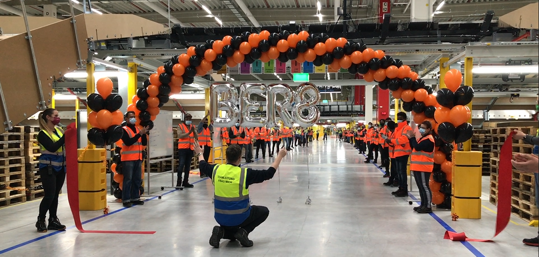 Mehrer Mitarbeiter mit Masken in einem Logistikzentrum: Alle klatschen, 2 Mitarbeiter halten einen Bogen mit schwarzen und orangenen Luftballons in die Höhe.