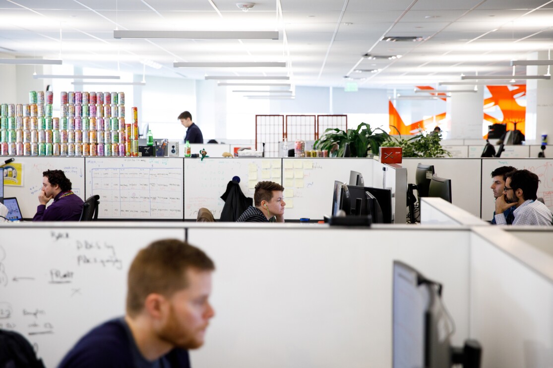 A view from within the Amazon office in Pittsburgh, Pennsylvania. Amazonians are sitting at desks divided by white boards. In the background, a wall of windows and some bright decor are seen.