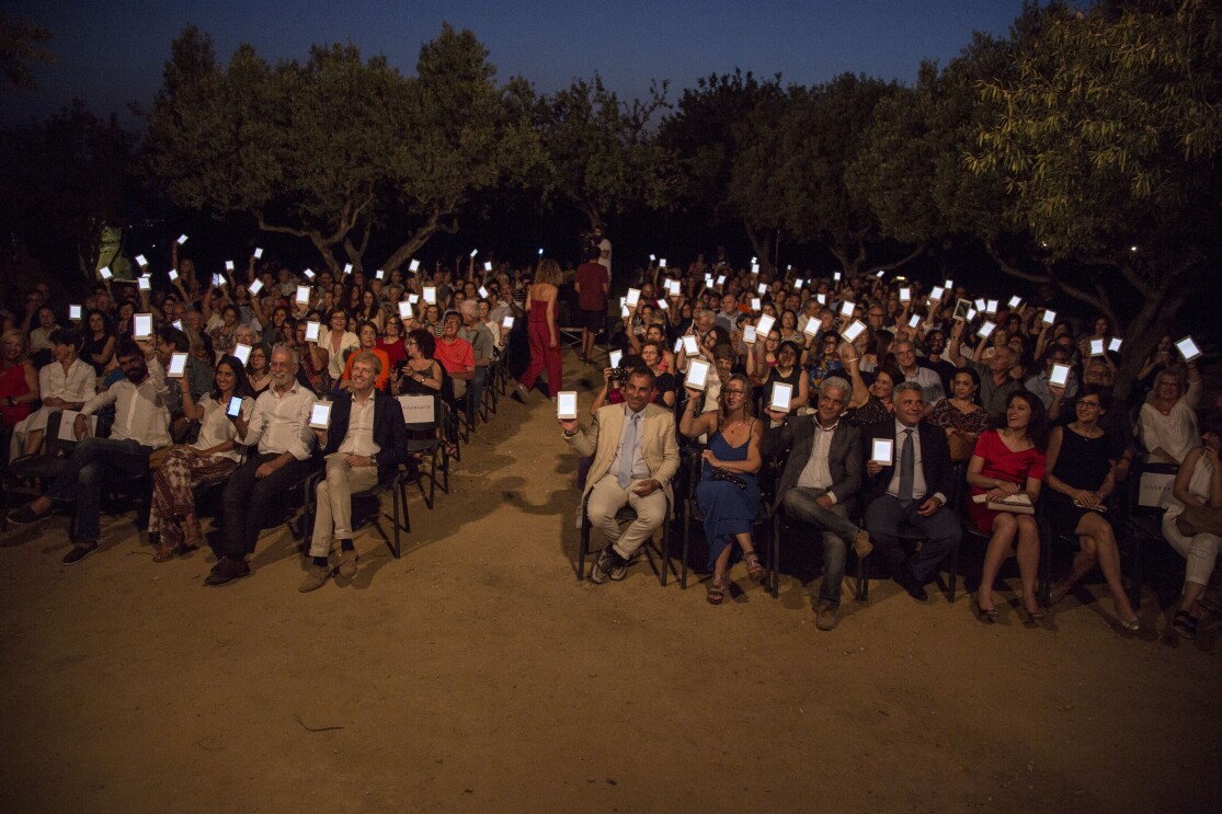 Platea di persone sedute nella Valle dei Templi di Agrigento durante un reading di Luigi Pirandello in occasione dei 150 della sua nascita. Gli spettatori reggono con una mano un Kindle PaperWhite, con lo schermo rivolto verso la camera. Sullo sfondo, degli alberi.