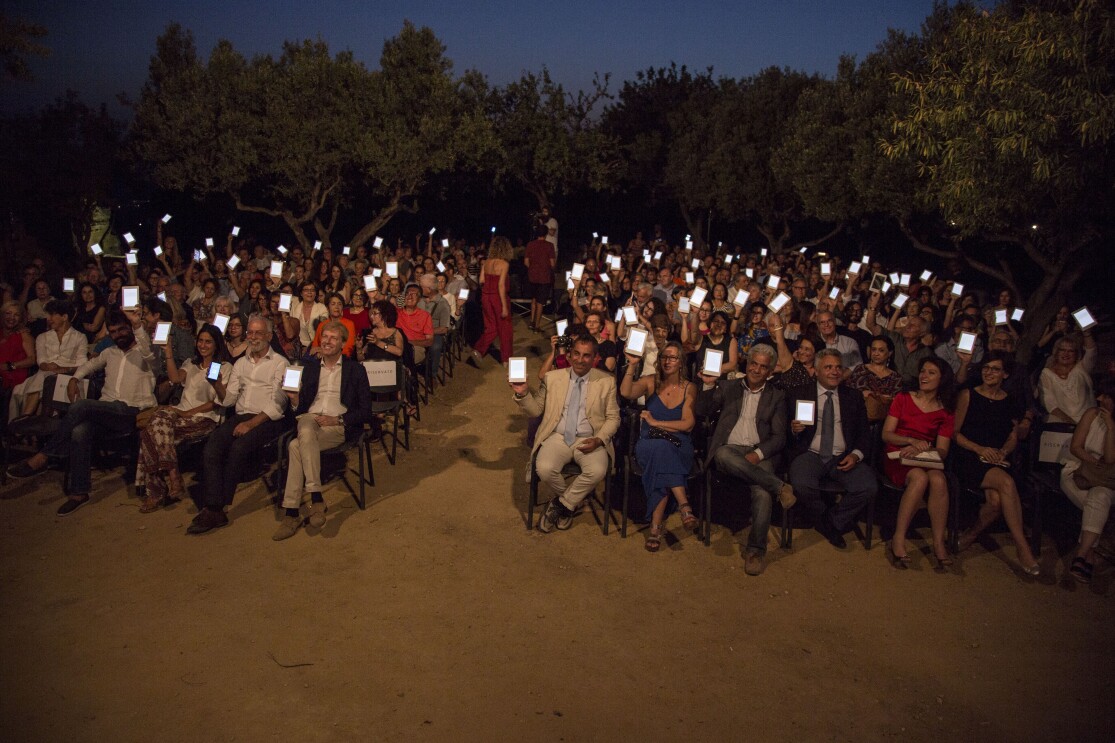 Platea di persone sedute nella Valle dei Templi di Agrigento durante un reading di Luigi Pirandello in occasione dei 150 della sua nascita. Gli spettatori reggono con una mano un Kindle PaperWhite, con lo schermo rivolto verso la camera. Sullo sfondo, degli alberi. 