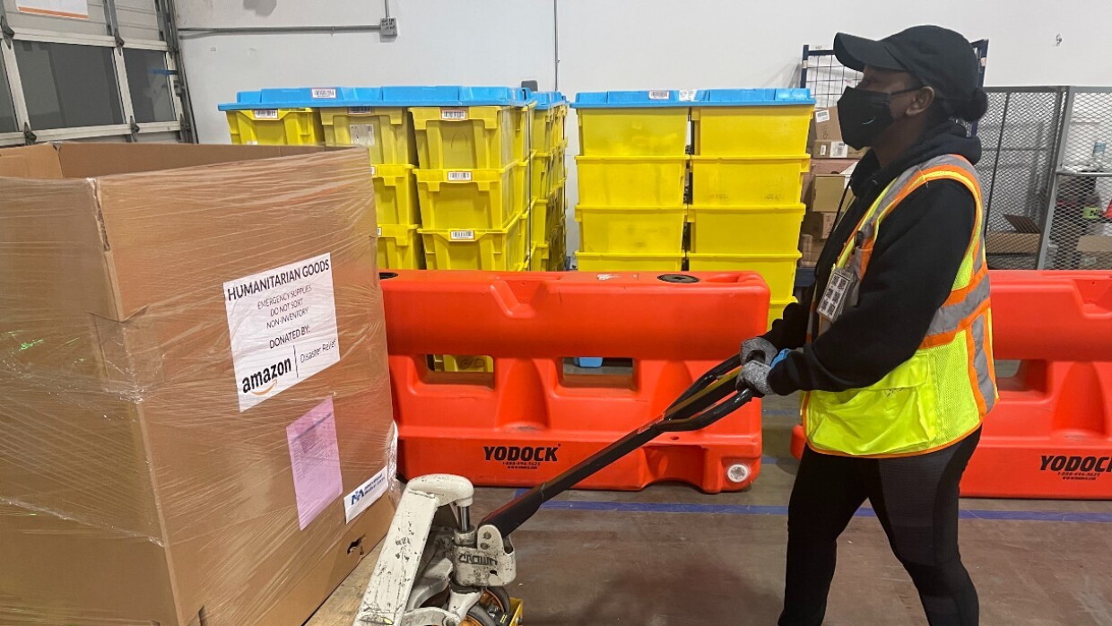 A woman wearing a yellow safety jacket and a face mask pushes a box that reads 'humantarian goods' with a pallet jack.
