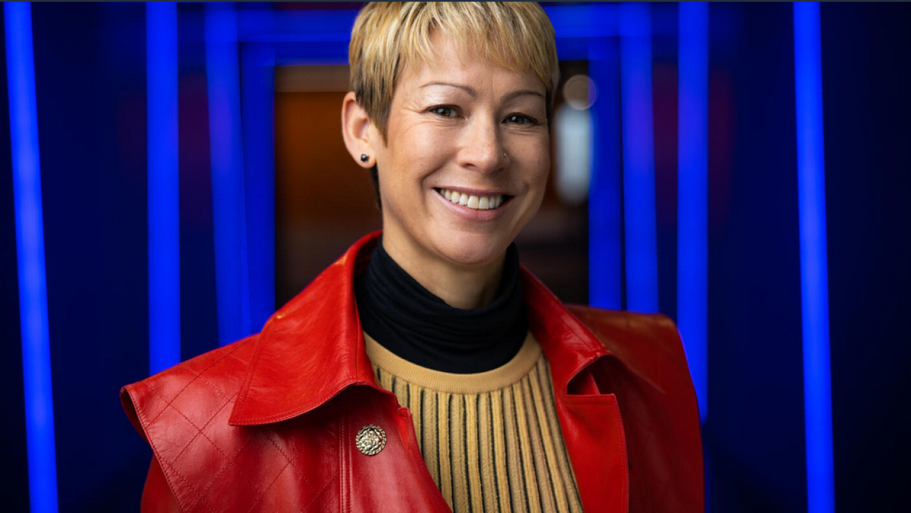 An image of a woman smiling for a headshot in a hallway of blue lights. She is wearing a red jacket and yellow sweater. 