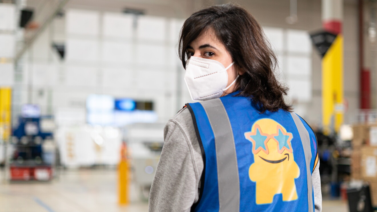 Silvia Mercado empezó hace dos años, de forma voluntaria, el proyecto Rodes màgiques. Silvia está de espladas pero con la cabeza girada mirando a cámara. De fondo hay un centro logístico con estanterías de color amarillo. Silvia lleva el pelo  negro por el hombro y escalonado. Su jerseiy es de color gris y lleva un chaleco de Amazon Box Star de color azul. 