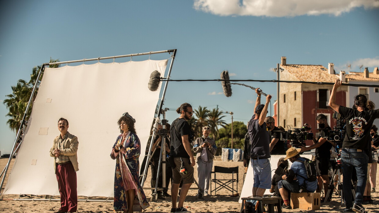 En un set de rodaje exterior, en la arena de la playa, aparecen dos actores, hombre y mujer vestidos de los años 60. Una pantalla blanca para aportiguar la luz. De fondo dos casa de pueblo marinero y, al lado de los actores, un equipo técnico de unas 10 personas con cámaras y micros. Todos van vestidos con ropa oscura. 