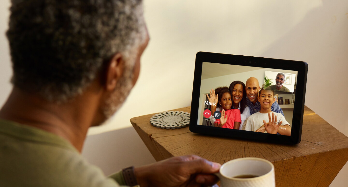 An Echo Show device on a wooden side table. On the screen, a family of four are smiling and waving on the screen. Facing the device is a man, holding a coffee cup. The screen shows his smiling face.
