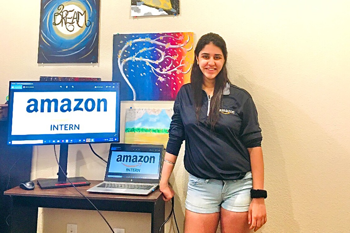 A woman stands next to a desk with a laptop and monitor displaying the words "Amazon intern."