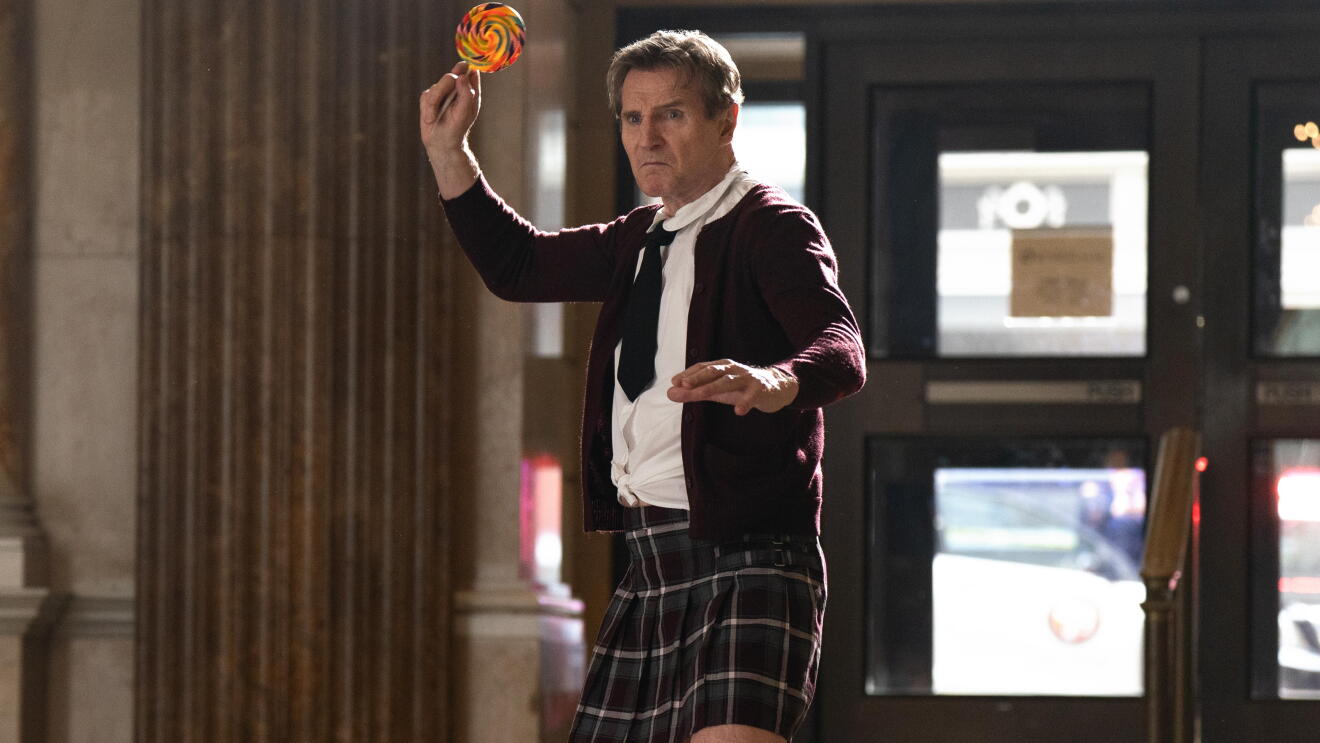 Liam Neeson wearing a girls school uniform and holding up candy